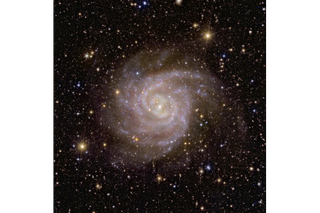 A pinkish, slightly hazy spiral galaxy in space, in front of many stars and more distant bright galaxies.